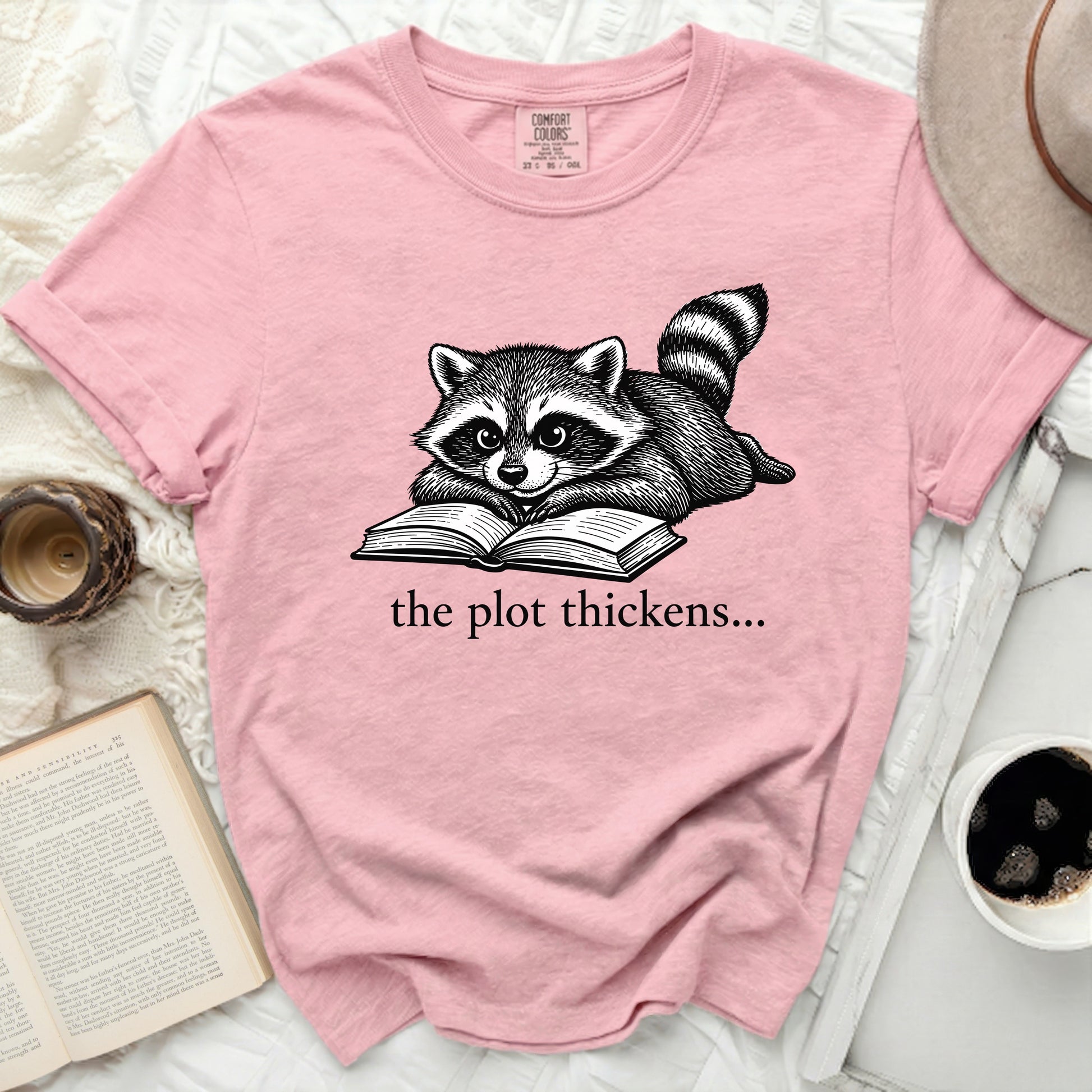 Pink t-shirt with a raccoon reading a book and text "the plot thickens...".