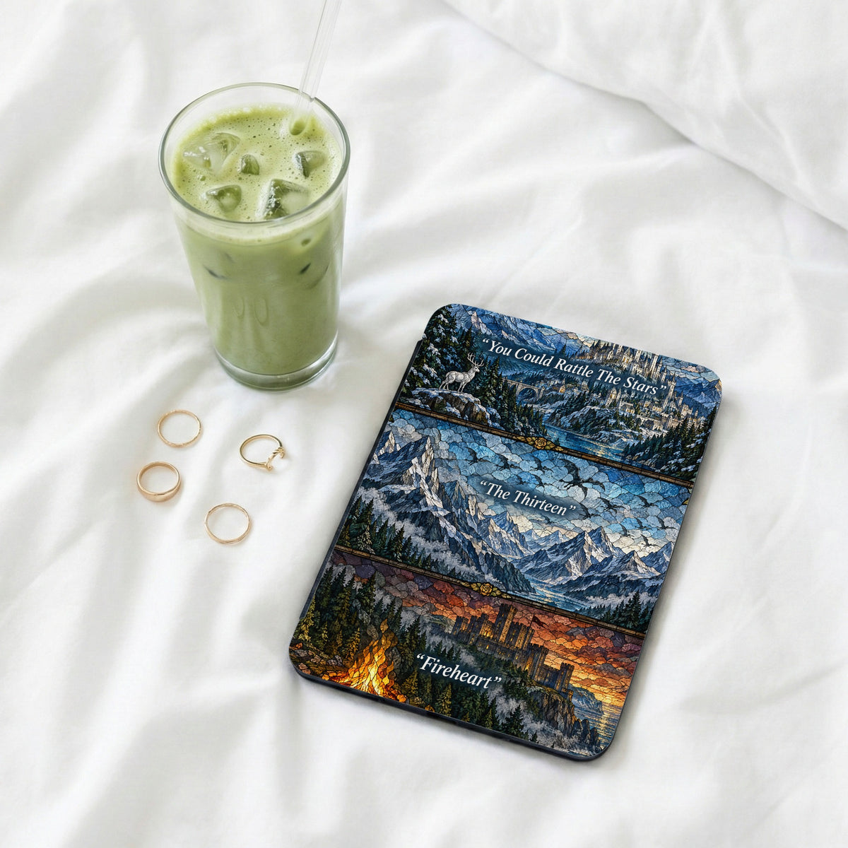 A Kindle reader with a stained-glass fantasy skin rests on white bedding next to an iced matcha latte and four gold rings.