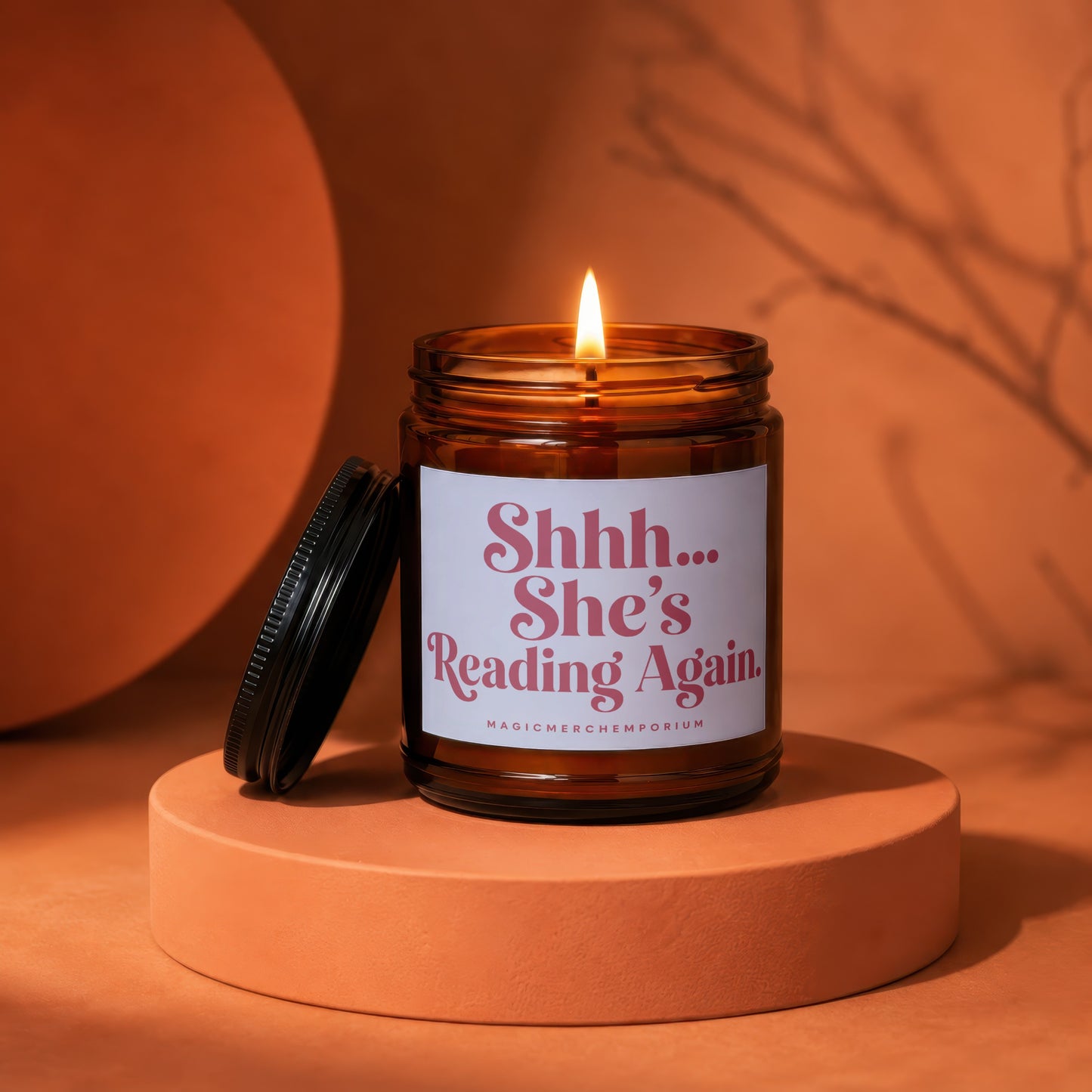 Amber glass jar candle with lit flame and the text "Shhh... She's Reading Again."