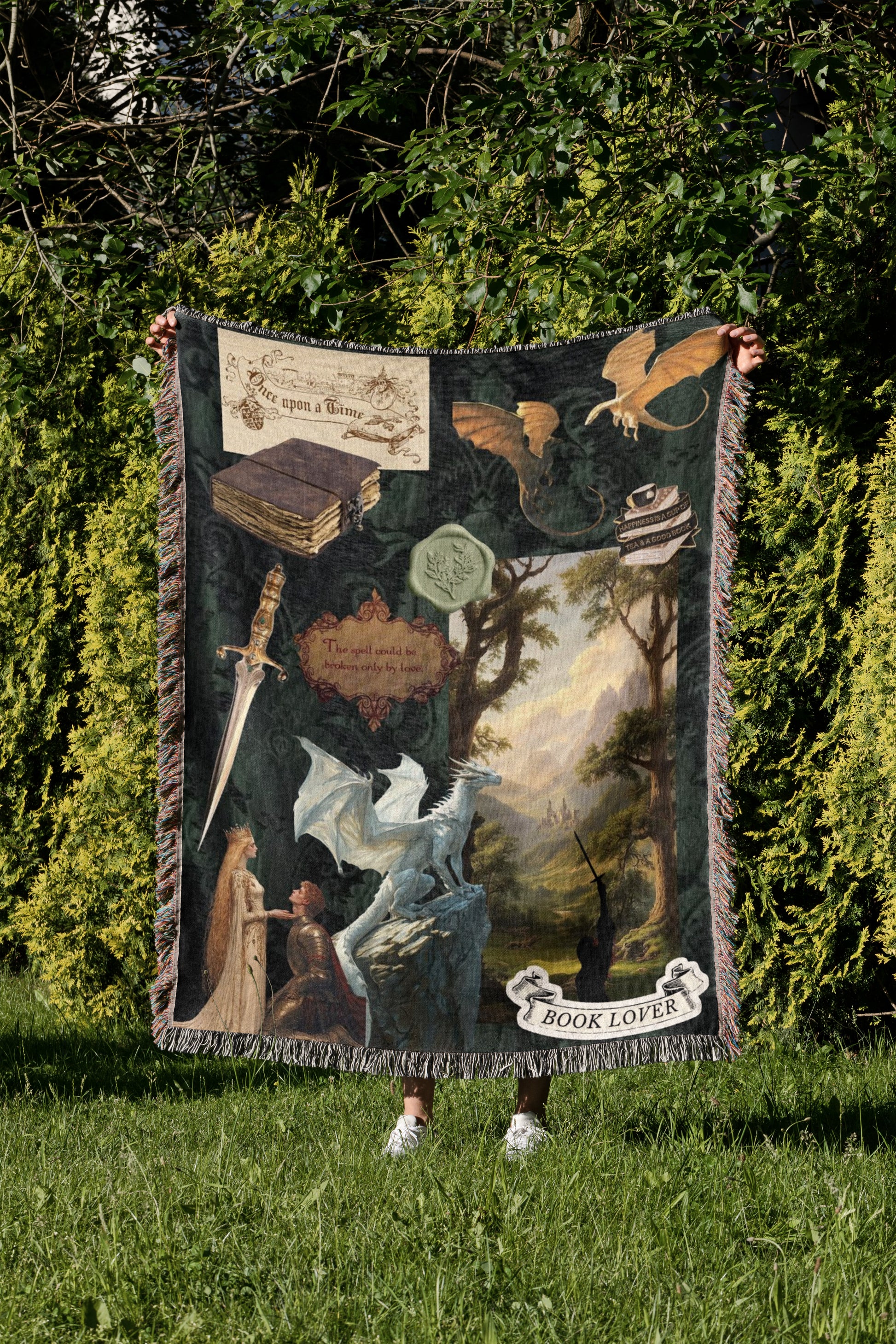 A person is standing in a grassy area, holding up a large blanket with a dragon and other fantasy-themed illustrations on it.