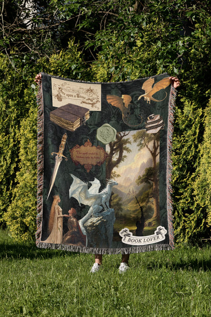 A person is standing in a grassy area, holding up a large blanket with a dragon and other fantasy-themed illustrations on it.