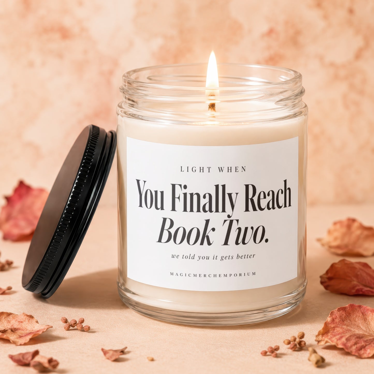 A lit candle in a glass jar with a black lid and label reading You Finally Reach Book Two.