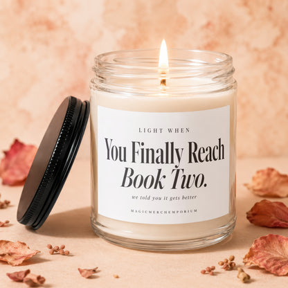 A lit candle in a glass jar with a black lid and label reading You Finally Reach Book Two.