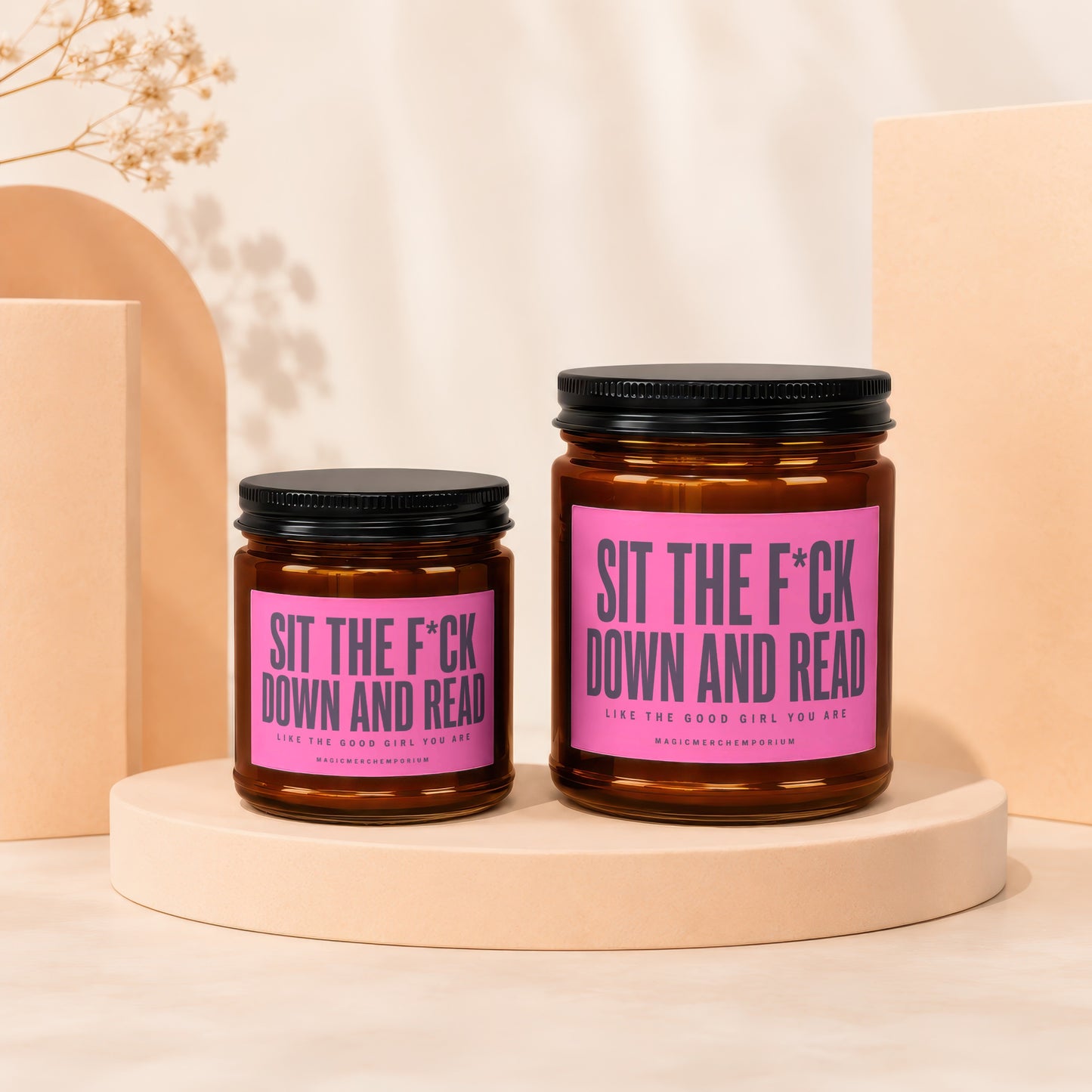 Two amber glass candles with black lids and pink labels reading "SIT THE F*CK DOWN AND READ".