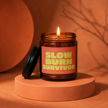 Amber jar candle with a lit flame sits on an orange pedestal with "Certified Slow Burn Survivor" text.