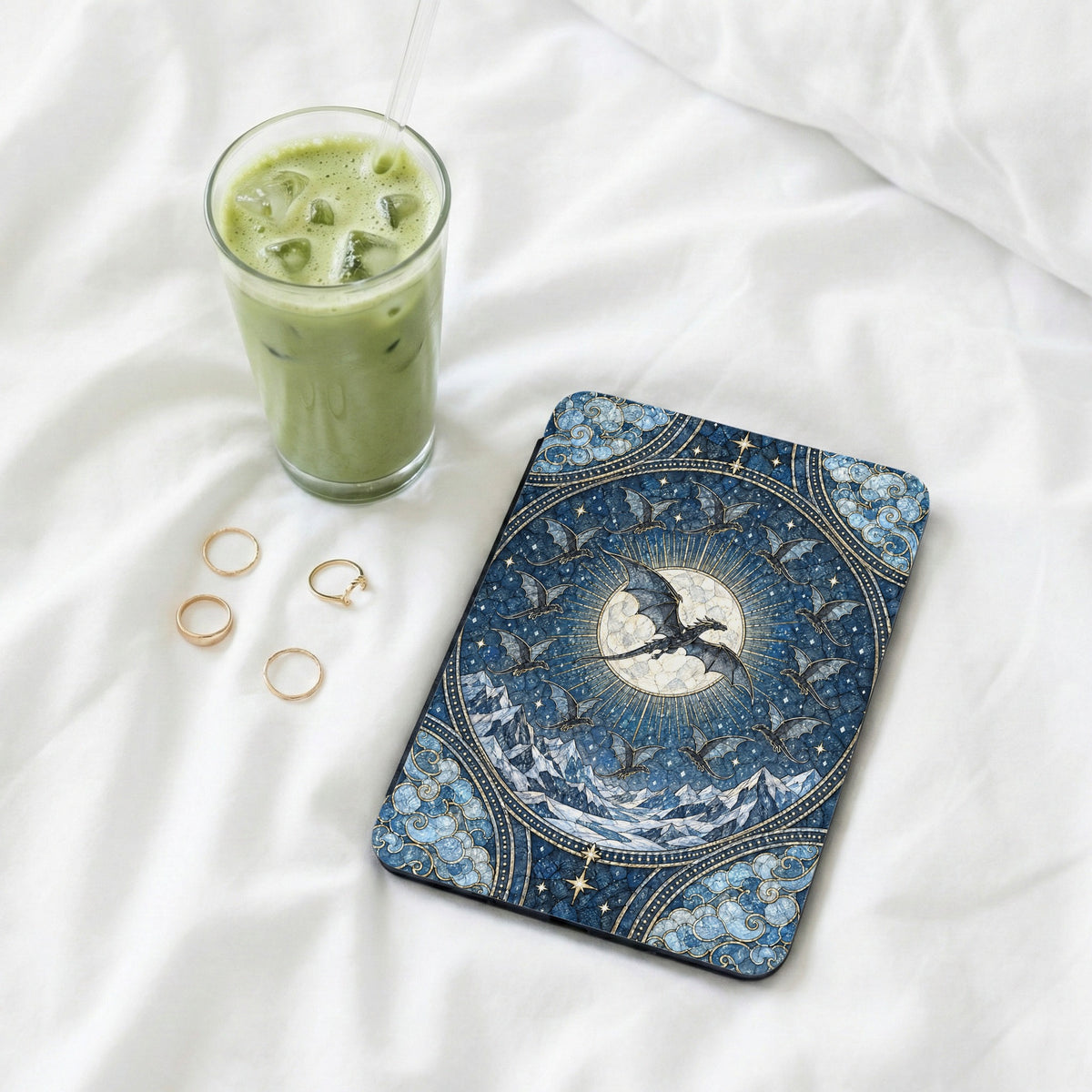 An e-reader in a blue fantasy dragon case sits on white bedsheets beside four gold rings and an iced green beverage.