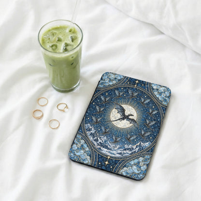 An e-reader in a blue fantasy dragon case sits on white bedsheets beside four gold rings and an iced green beverage.