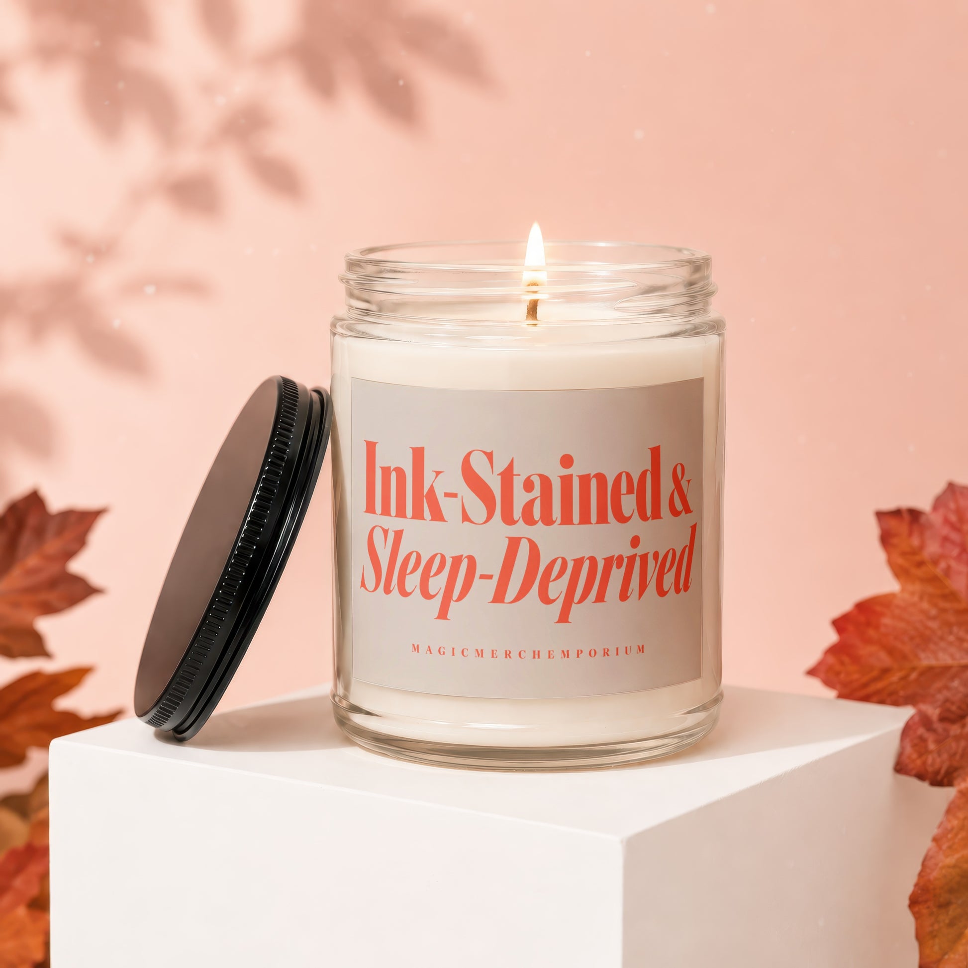 Glass candle with "Ink-Stained & Sleep-Deprived" label and lit wick.