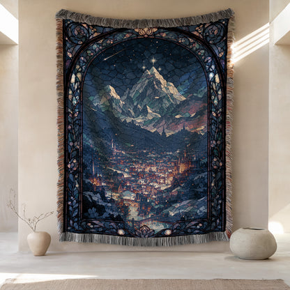 a large tapestry hanging on a wall, depicting a mountainous landscape with a city in the background. The tapestry has a detailed and intricate design, showcasing the craftsmanship and artistry of the creator. The tapestry is framed by a decorative border, adding to its visual appeal. The tapestry is placed on a wall, which is adjacent to a small vase and a potted plant, creating a harmonious and balanced composition in the room.