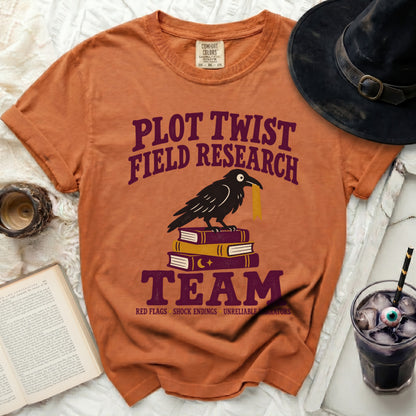 Orange t-shirt with "Plot Twist Field Research Team" text, crow on books.