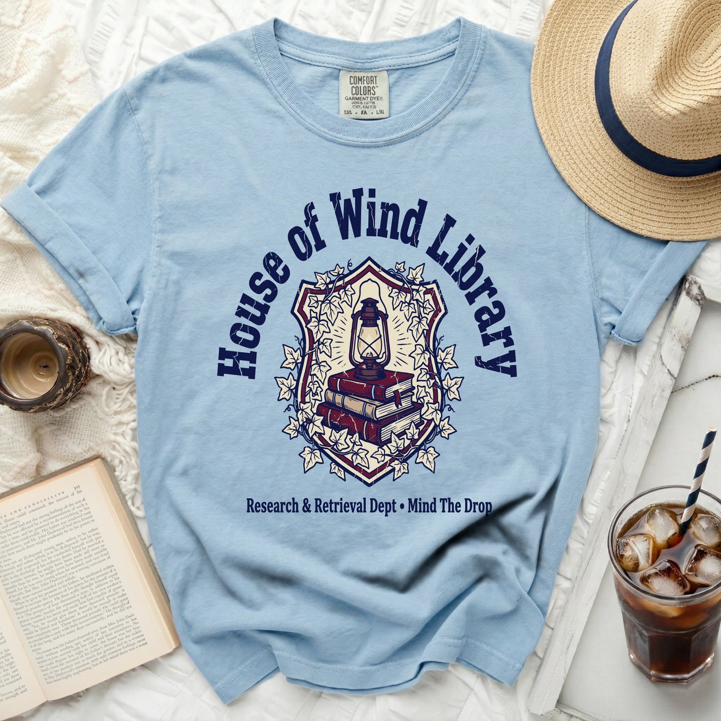 Light blue t-shirt with "House of Wind Library" crest and book design.