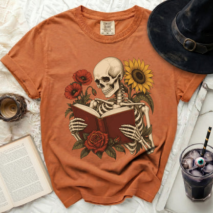Skeleton reading a book surrounded by poppies and a sunflower on an orange t-shirt.