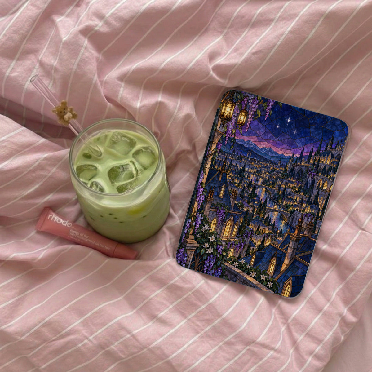 Phone case with stained glass castle scene, iced matcha latte, and lip balm on pink striped fabric.