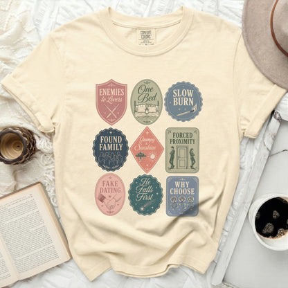 Cream t-shirt with nine circular book trope badges in pastel colors.
