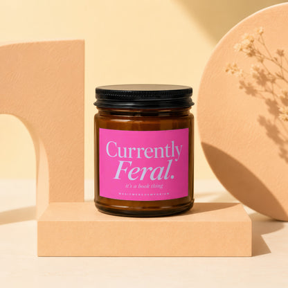 Amber jar candle with pink label that reads "Currently Feral. it's a book thing".
