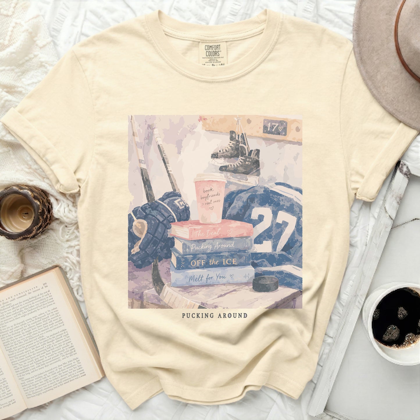 Cream t-shirt with stacked books, hockey gear, and coffee cup.
