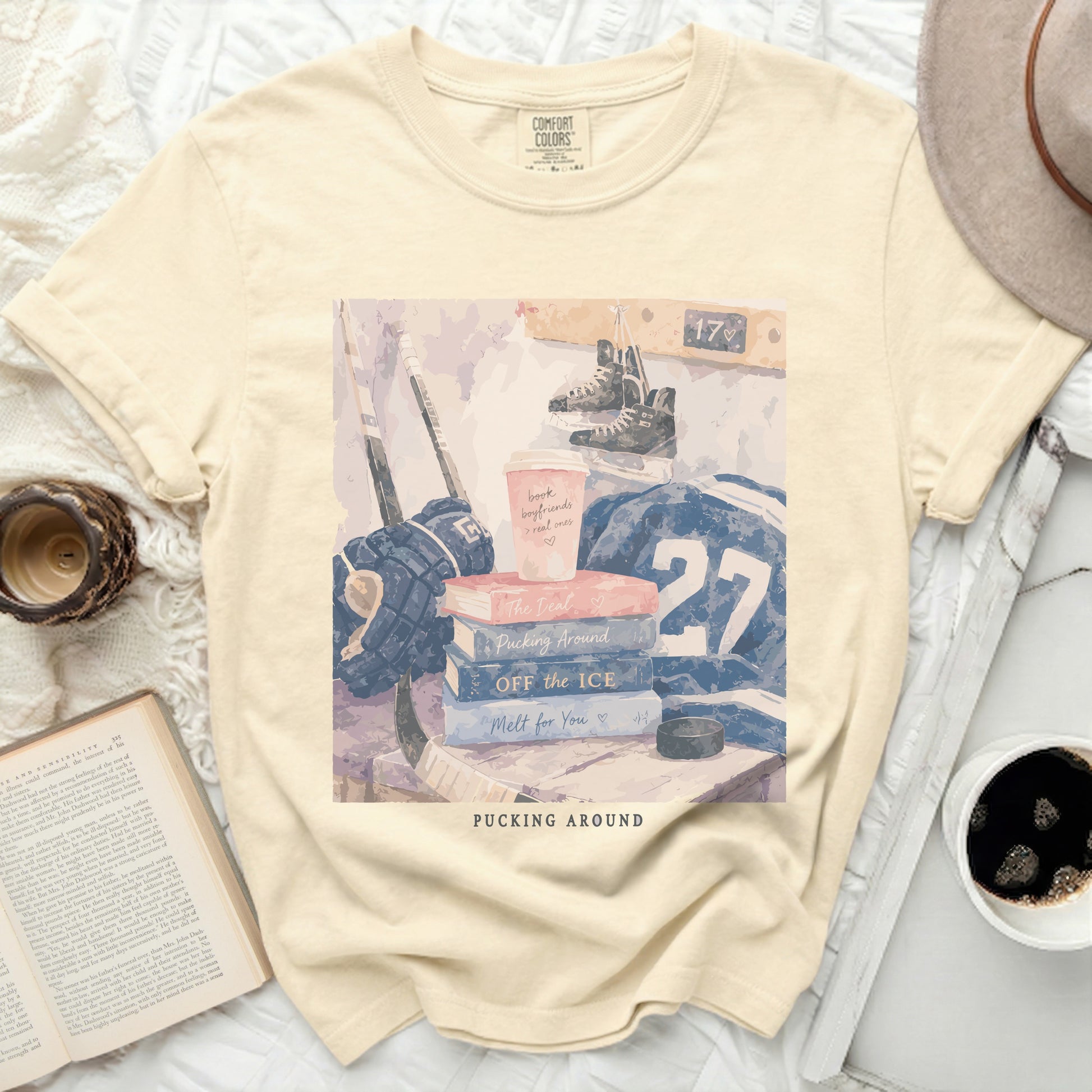 Cream t-shirt with stacked books, hockey gear, and coffee cup.