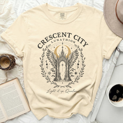 A beige Comfort Colors t-shirt features a black and gold graphic of winged archway, crescent moon, and the text Crescent City Lunathion Light it up, Quinlan.