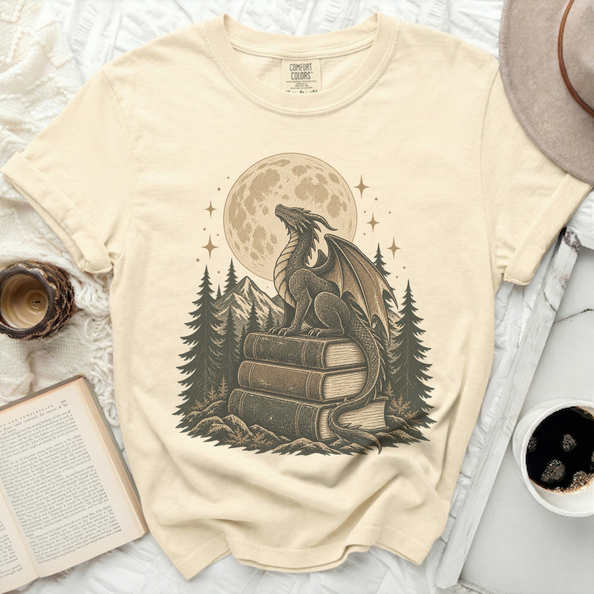 Dragon on books in front of moon and pine trees graphic on cream t-shirt.