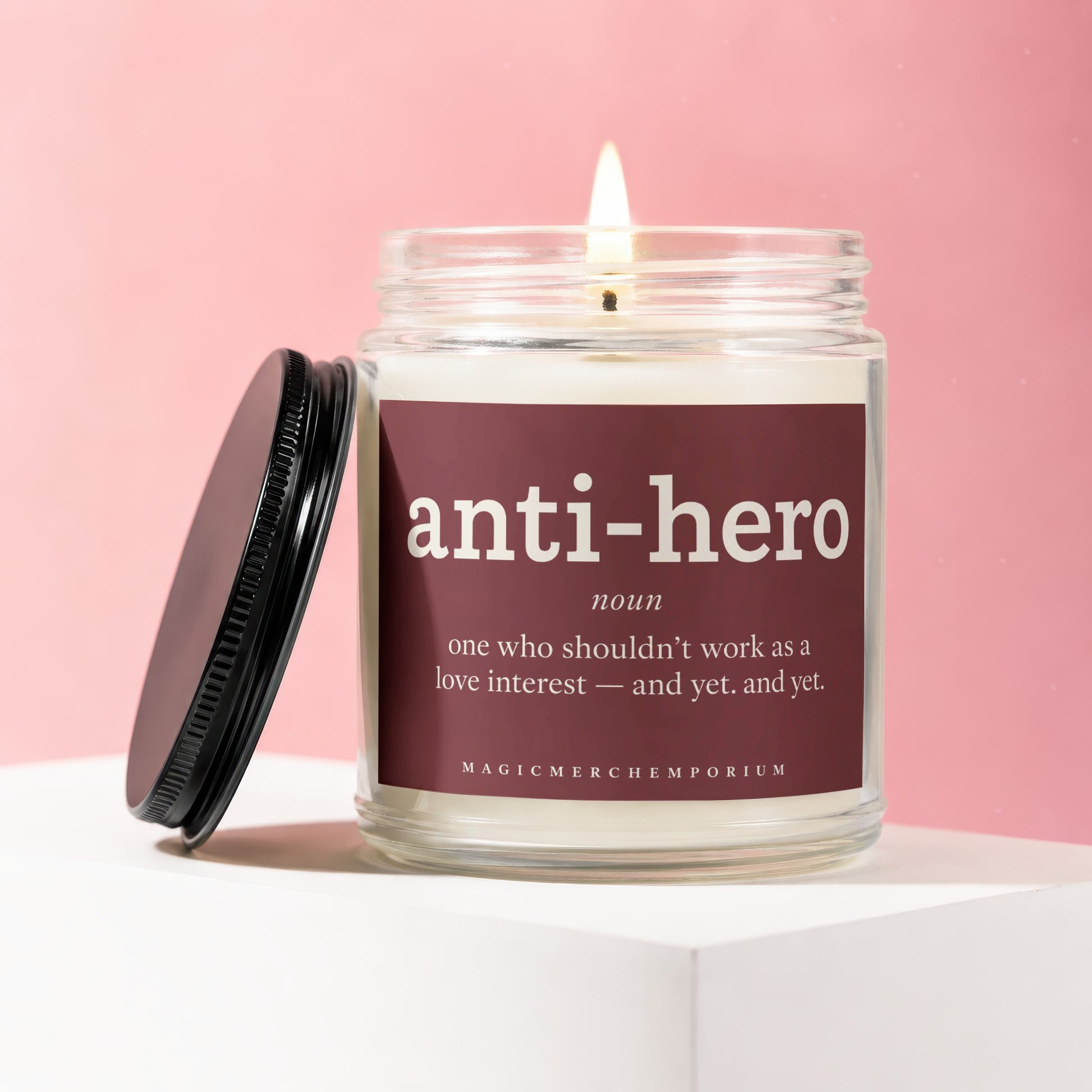 Glass jar candle with lit wick, featuring "anti-hero" definition on maroon label.