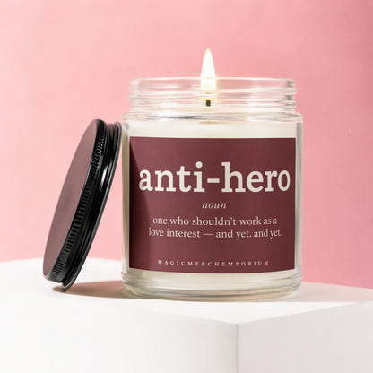 Glass jar candle with lit wick, featuring "anti-hero" definition on maroon label.