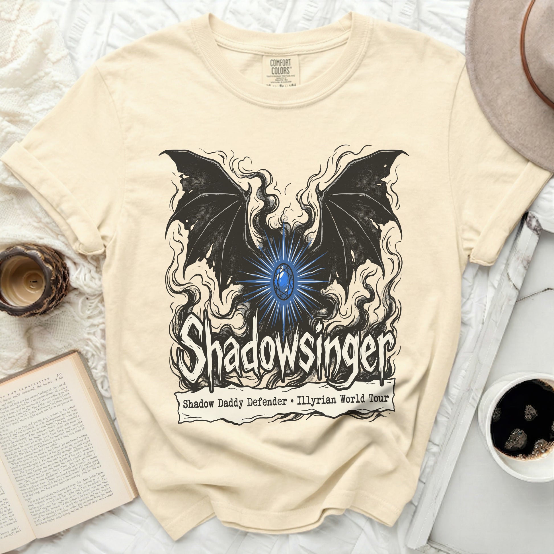 Cream t-shirt with black graphic of wings, a blue gem, and text "Shadowsinger".