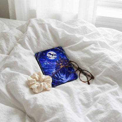 Kindle reader with a blue stained-glass castle case rests on a white bed alongside glasses and a scrunchie.