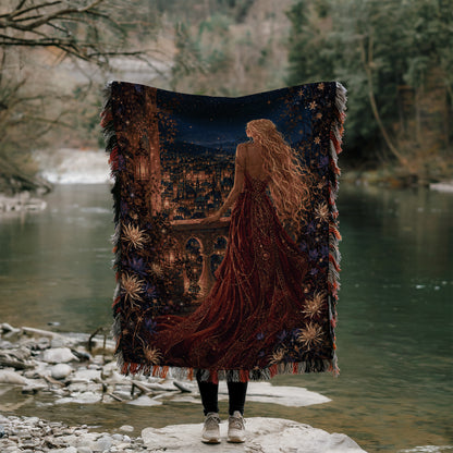 A person stands on a rock, holding a large tapestry that depicts a woman with long, flowing hair and a red dress, gazing out at a river or lake.