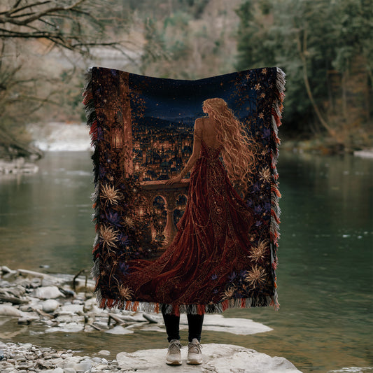 A person stands on a rock, holding a large tapestry that depicts a woman with long, flowing hair and a red dress, gazing out at a river or lake.