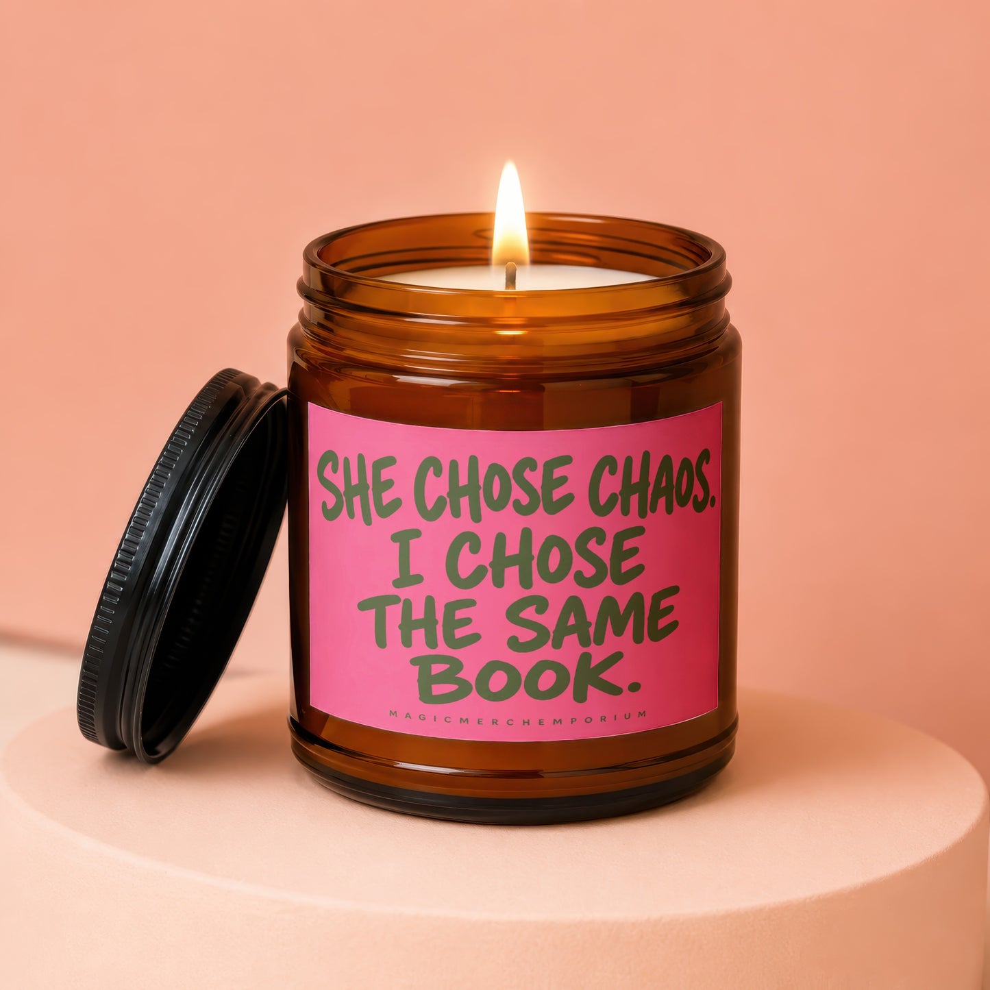 Amber jar candle with pink label reading "She chose chaos. I chose the same book."