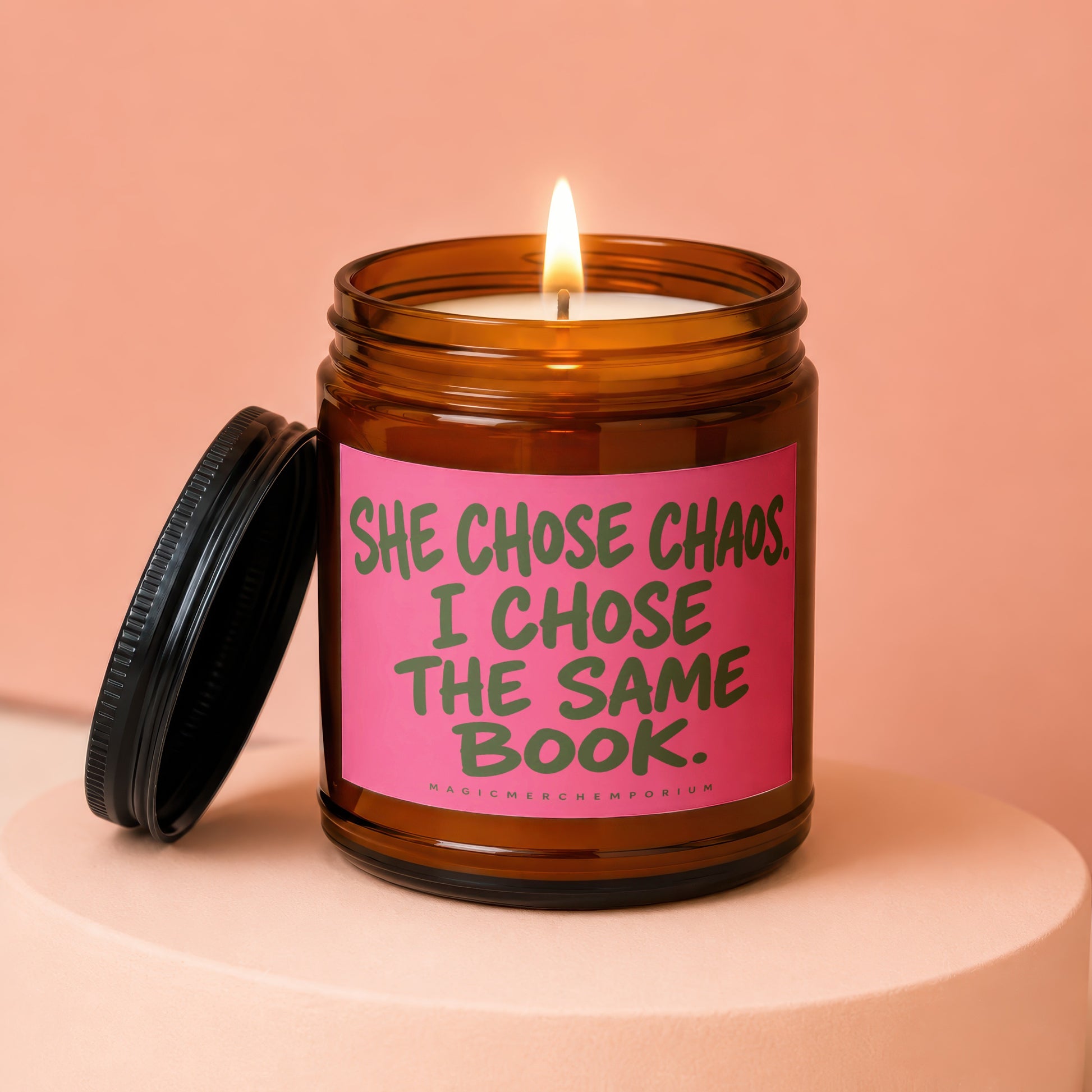 Amber jar candle with pink label reading "She chose chaos. I chose the same book."