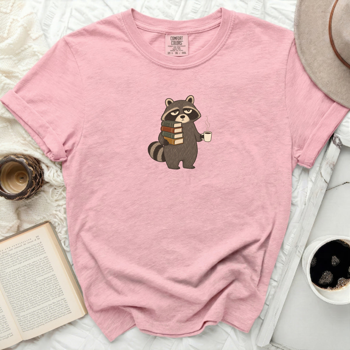 Pink t-shirt with a cartoon raccoon holding books and a coffee mug.