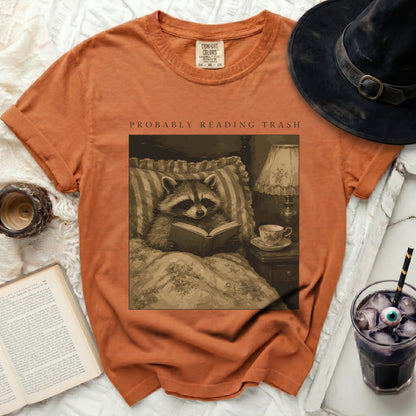 Orange t-shirt with graphic of raccoon reading in bed, "PROBABLY READING TRASH" text.