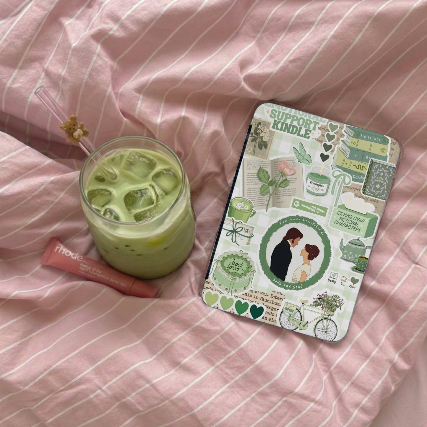 Green iced matcha drink and a sticker-covered Kindle on a pink striped blanket.