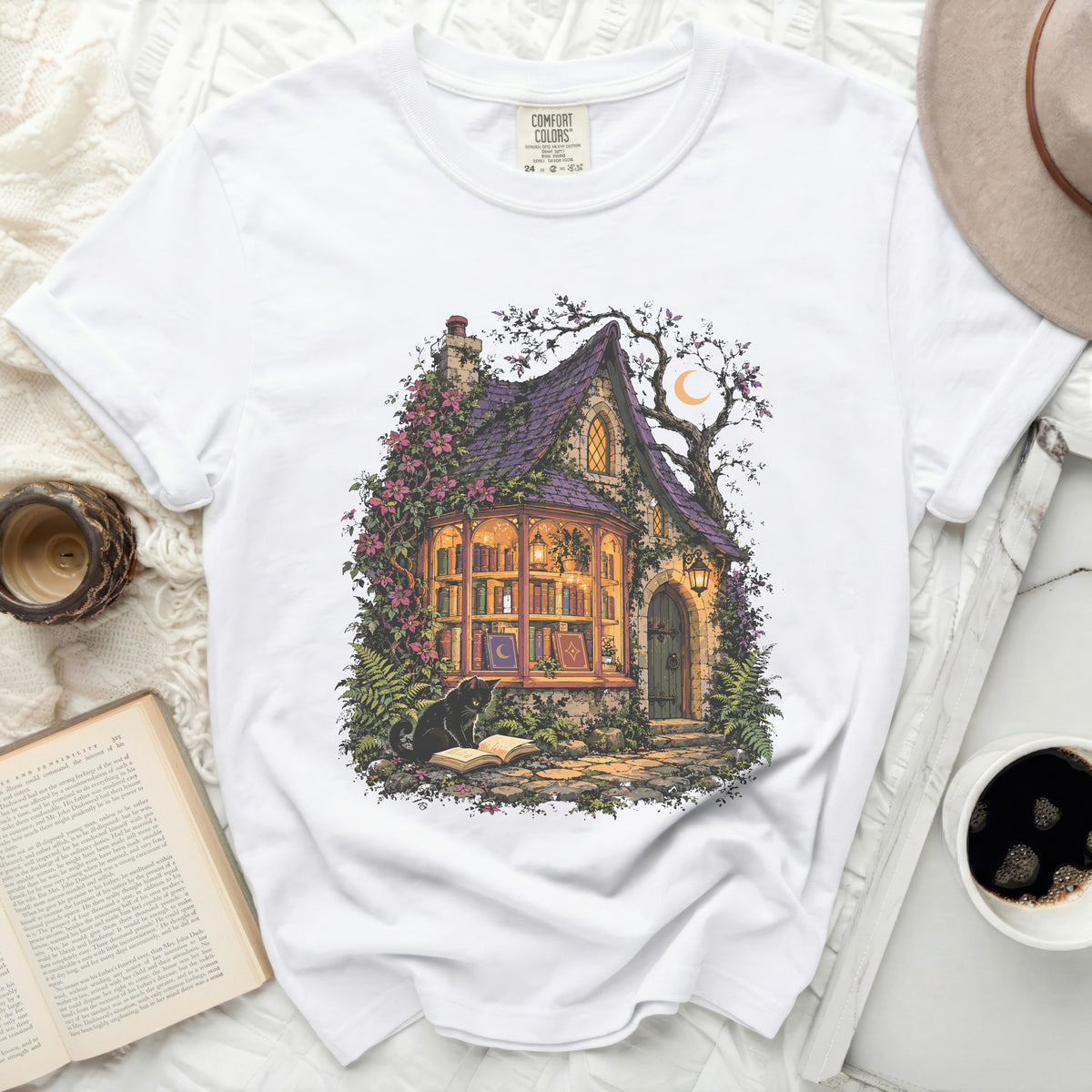 White t-shirt with graphic of a witch's cottage, black cat, and books.
