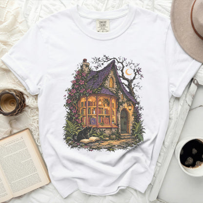 White t-shirt with graphic of a witch's cottage, black cat, and books.