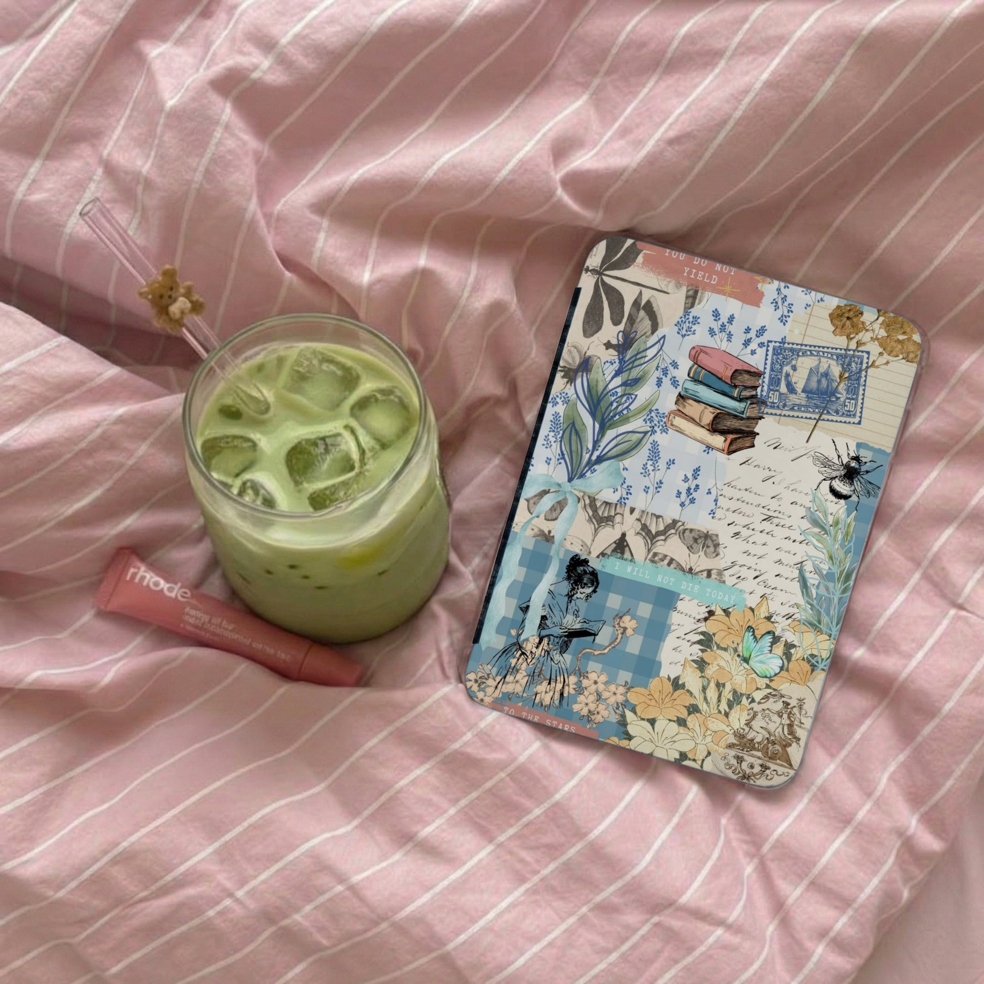 Iced matcha latte, Rhode lip balm, and a journal with collage art on pink striped fabric.
