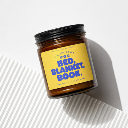Amber glass candle with yellow label reading "Bed, Blanket, Book."