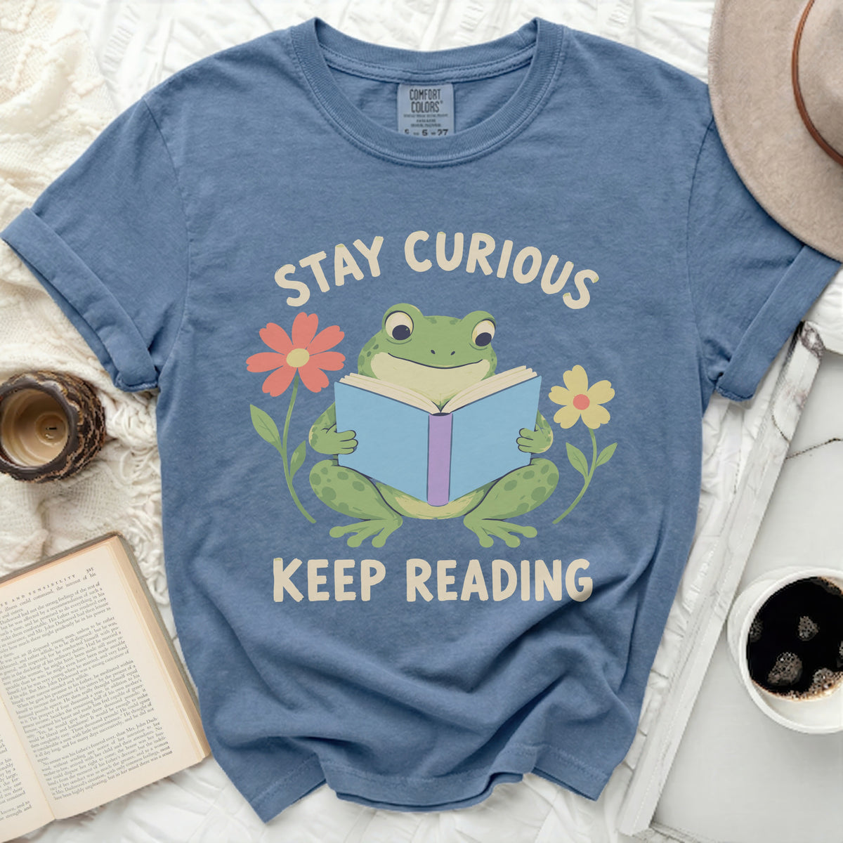 A blue t-shirt printed with a green frog reading a book, surrounded by flowers and the text Stay Curious Keep Reading.