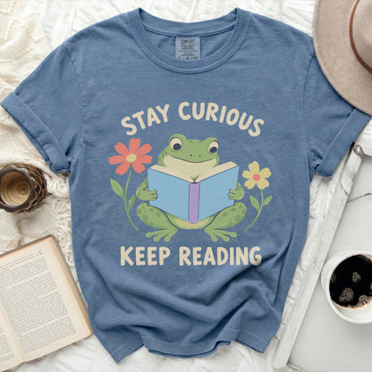 A blue t-shirt printed with a green frog reading a book, surrounded by flowers and the text Stay Curious Keep Reading.