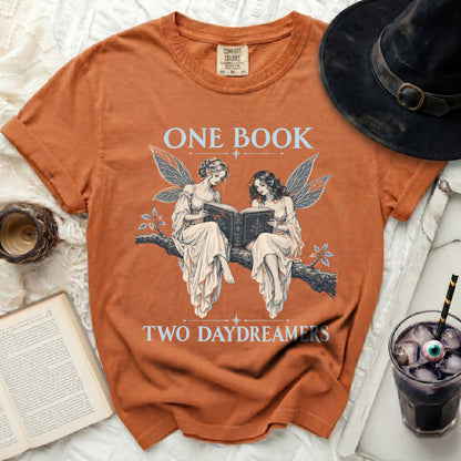 Orange t-shirt with two winged fairies reading a book, text "ONE BOOK TWO DAYDREAMERS".