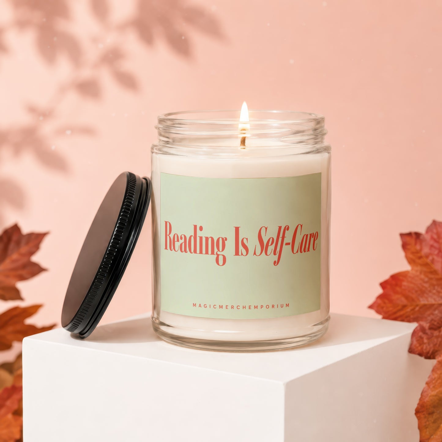 Lit candle with "Reading Is Self-Care" text, lid off, on a white cube.