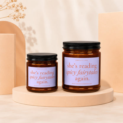 Two amber jar candles with black lids on display pedestals, text reads "she's reading spicy fairytales again."