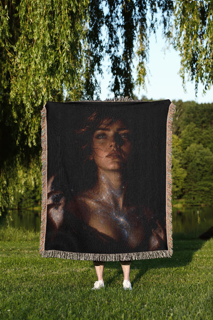 A person stands in a grassy field, holding a large square blanket featuring a portrait of a woman with long, dark hair.