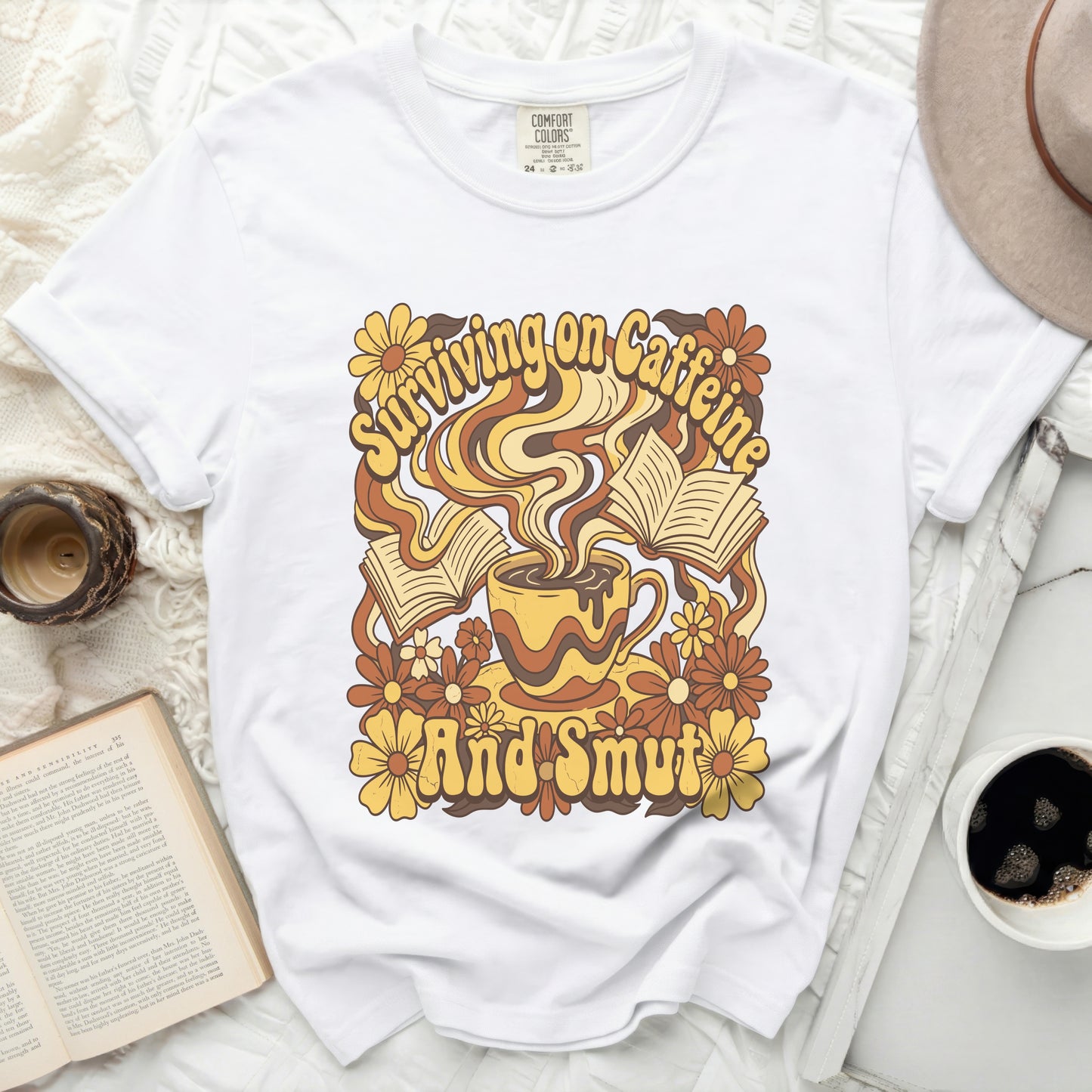 White t-shirt with retro graphic of coffee cup, books, and flowers, text "Surviving on Caffeine And Smut".