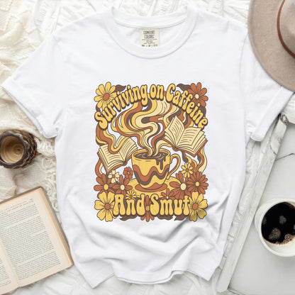 White t-shirt with retro graphic of coffee cup, books, and flowers, text "Surviving on Caffeine And Smut".