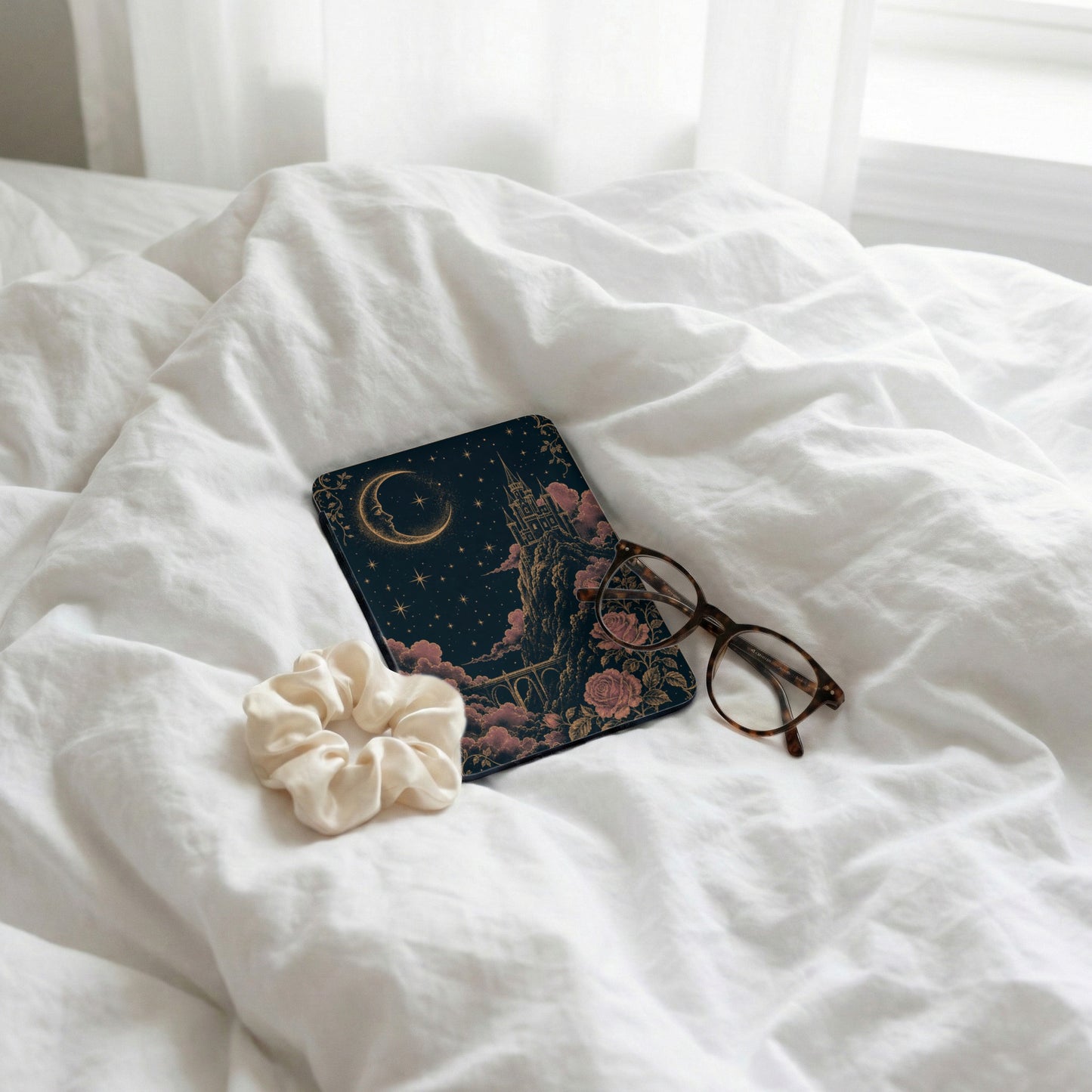 A celestial castle design on a dark cover rests on white bedsheets with eyeglasses and scrunchie.