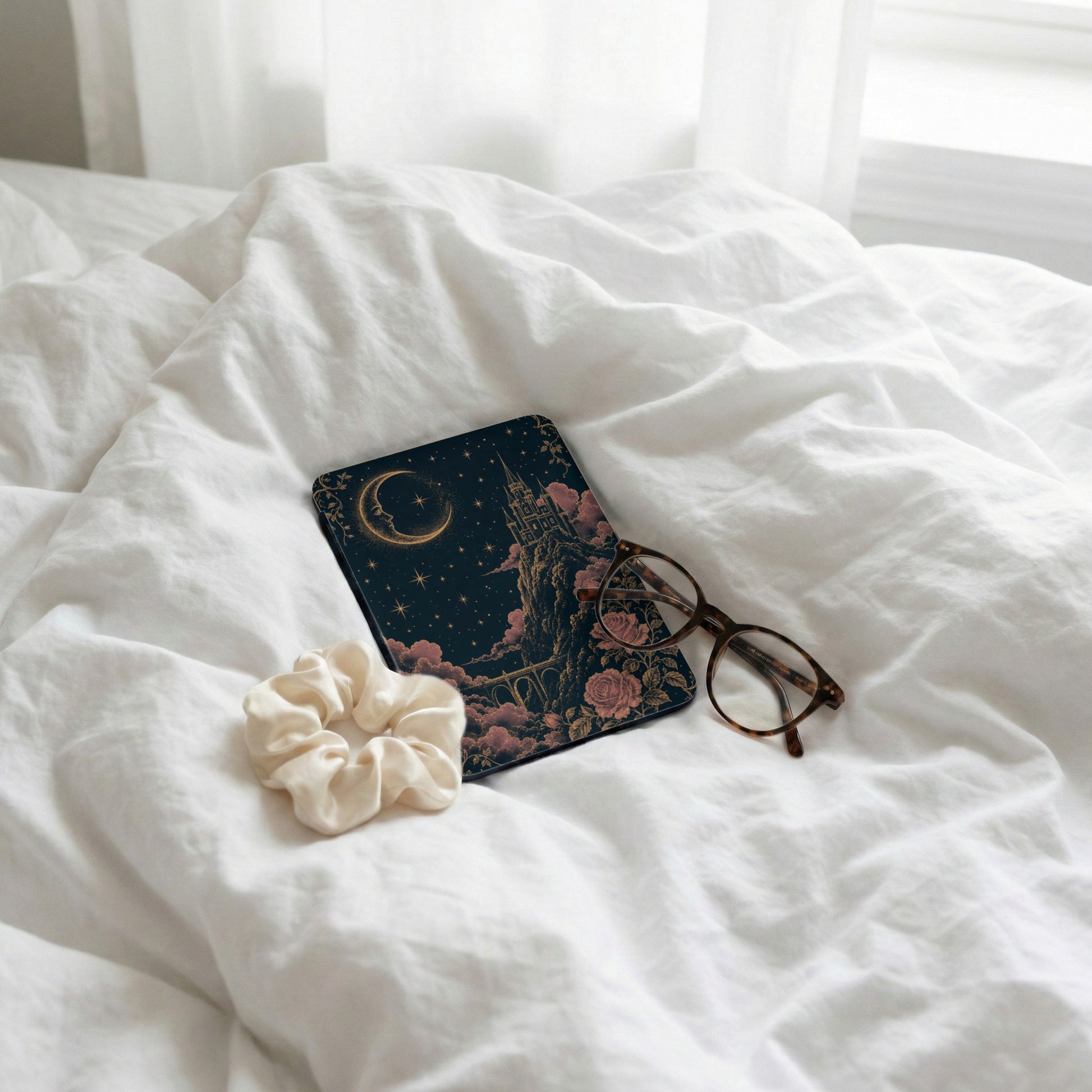 A celestial castle design on a dark cover rests on white bedsheets with eyeglasses and scrunchie.