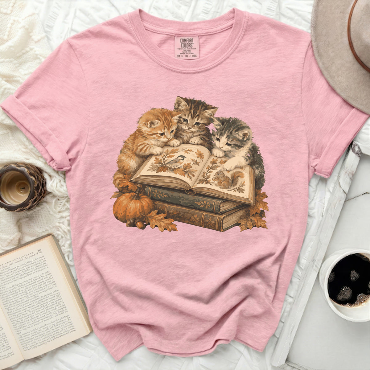 Pink t-shirt with a graphic of three kittens reading an open book with autumn leaves and a pumpkin.