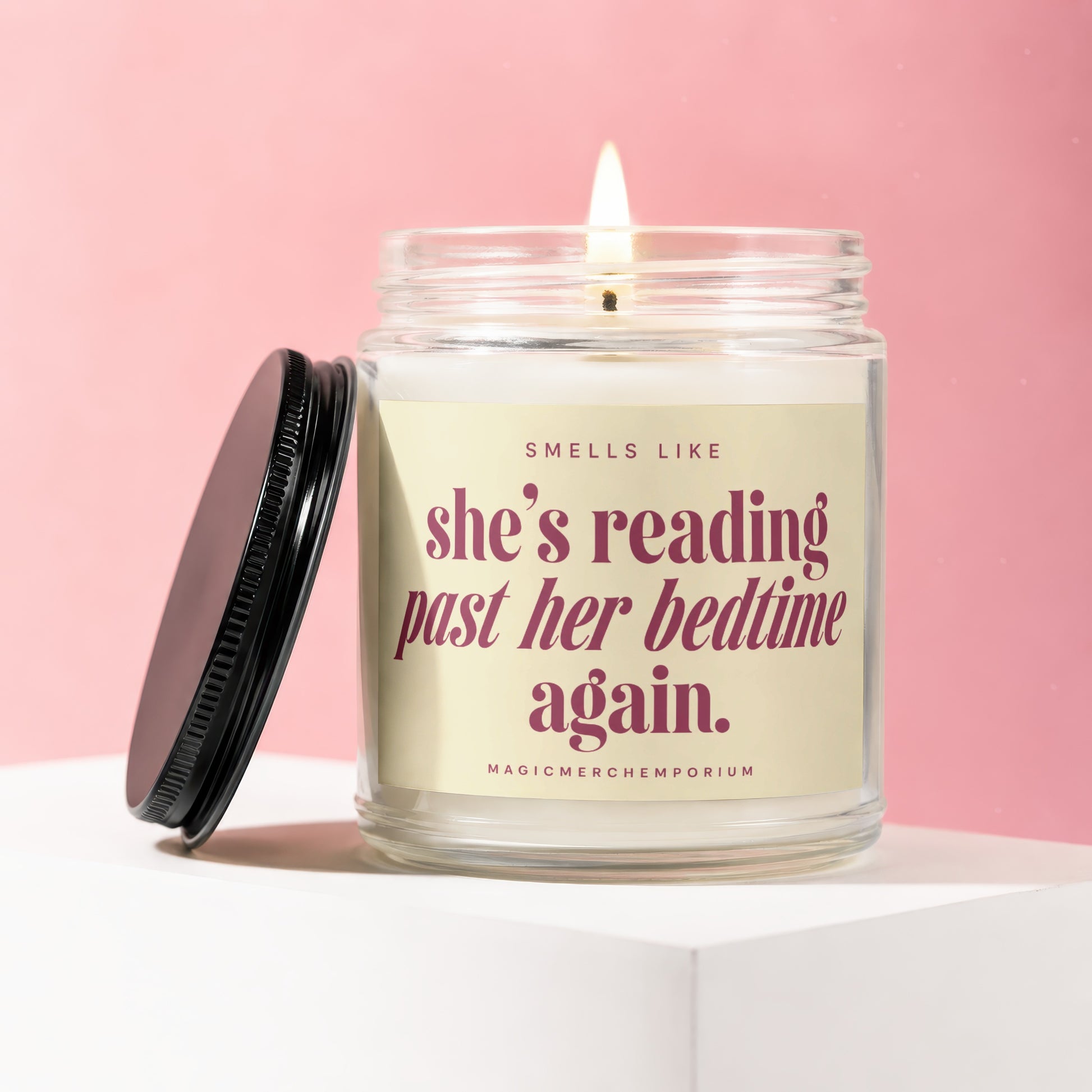 Glass candle with lit wick and black lid, says "SMELLS LIKE she's reading past her bedtime again. MAGICMERCH..."